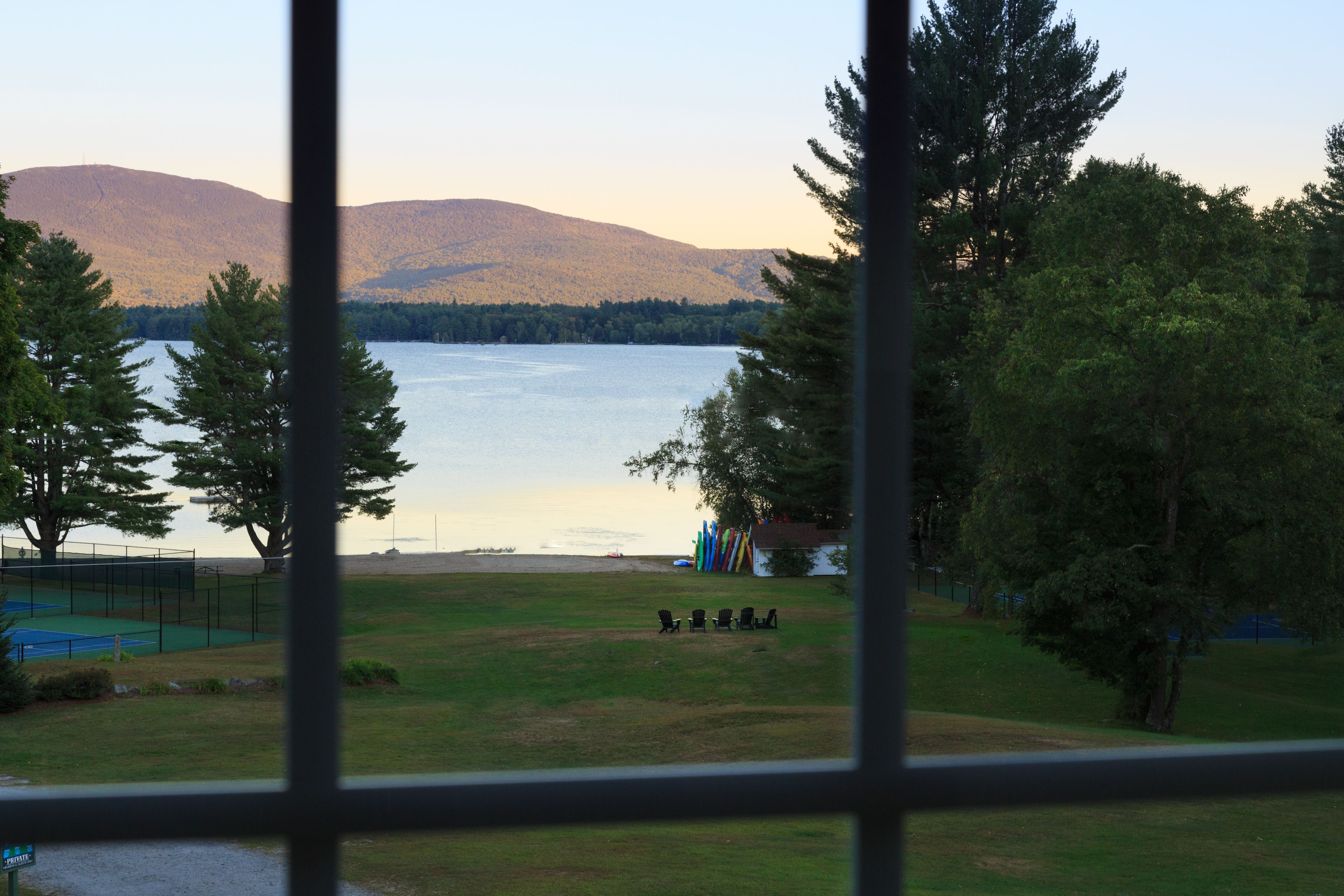 A view of a lake with mountains in the background, a grassy area with chairs, and trees.