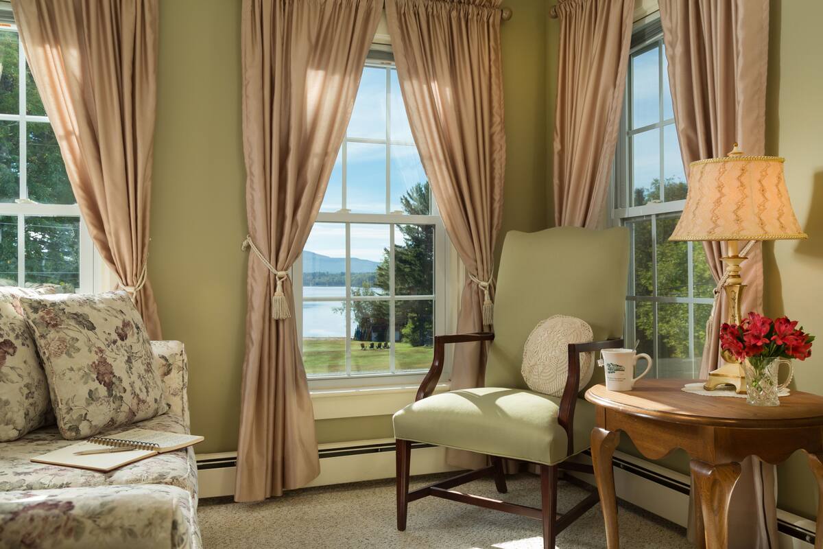 A cozy room with a floral sofa, a wooden chair, a small table with a lamp and flowers, and large windows with curtains.