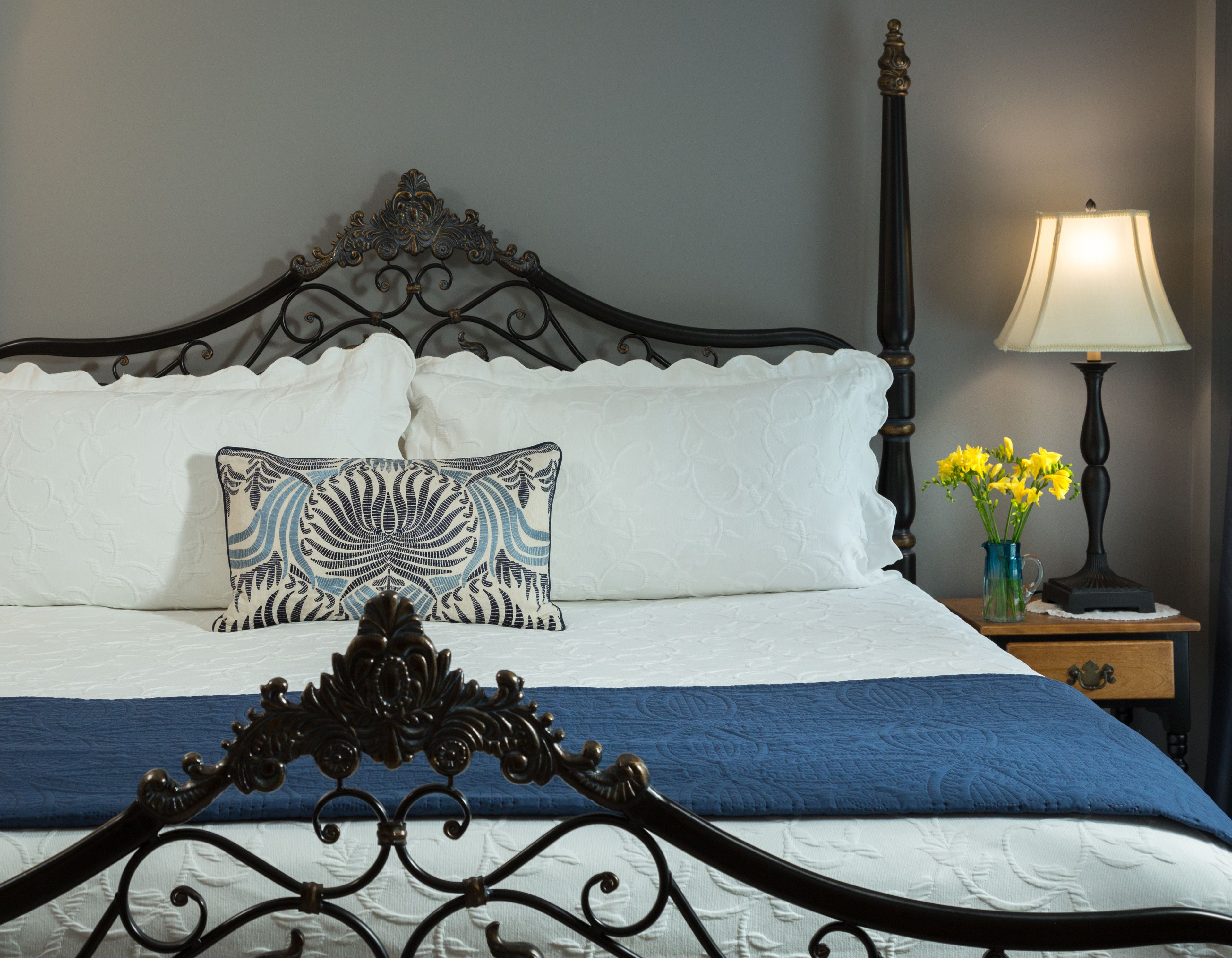 Un lit bien fait, agrémenté d’une tête de lit décorative, d’une couverture bleue et blanche et d’un coussin à motifs. On y trouve une table de chevet avec une lampe et un vase rempli de fleurs jaunes.