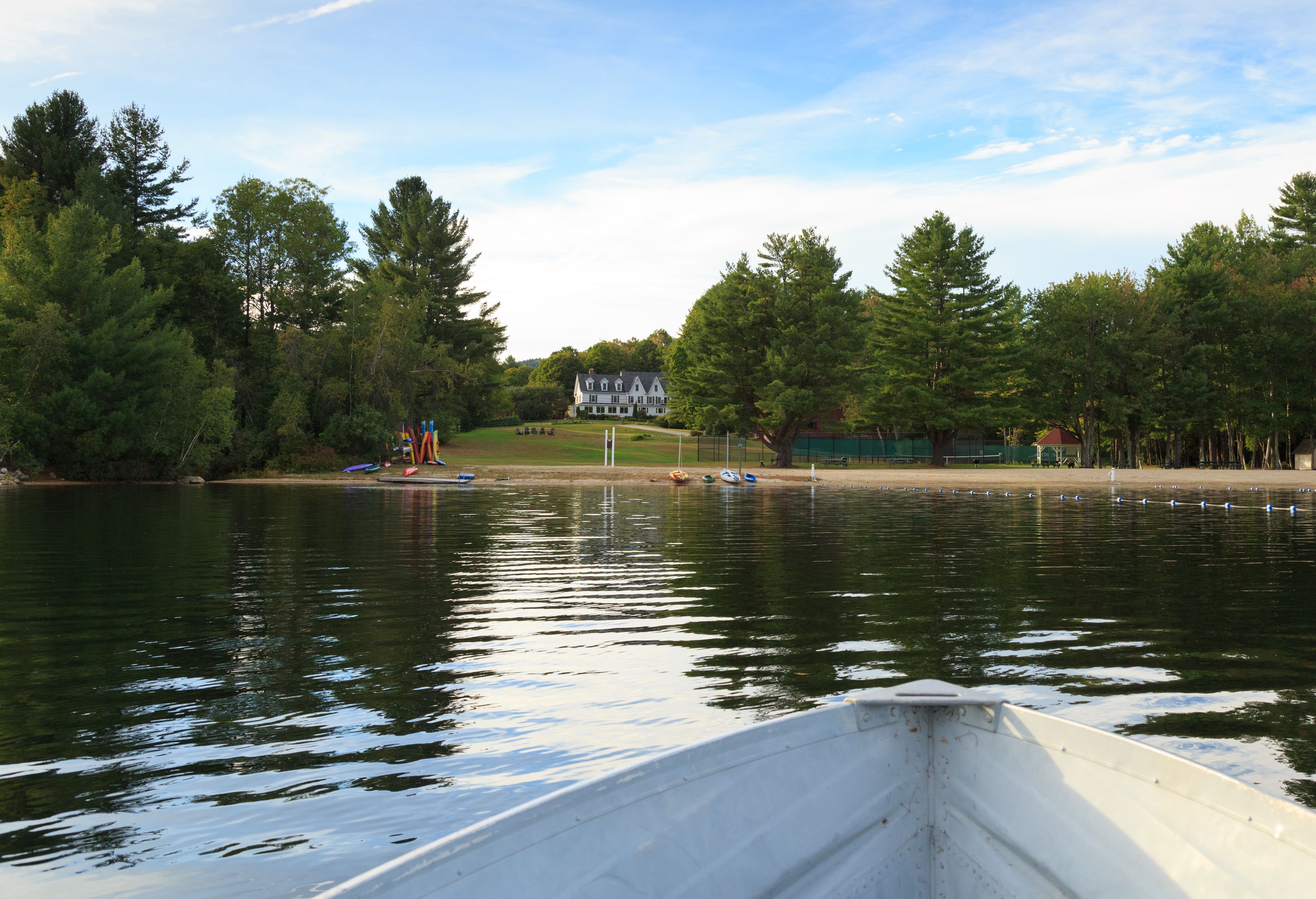 A lake with a boat in the foreground, a large house in the background, and a forested shoreline.