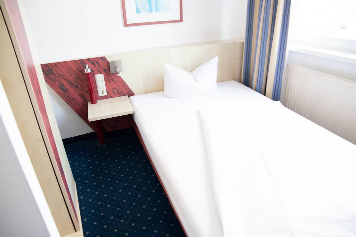 A hotel room with a bed, a nightstand, a red telephone, and a window with curtains.