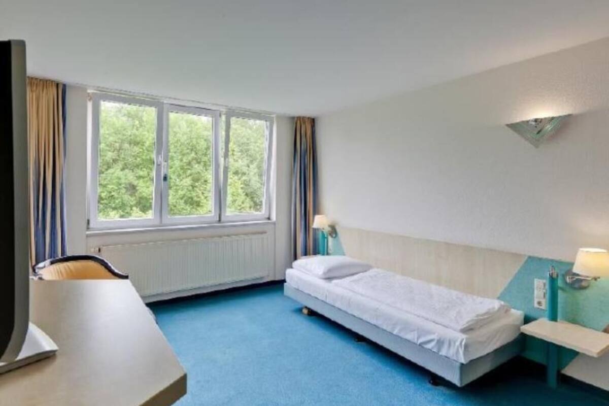 A hotel room with a bed, a desk, a chair, a window with curtains, and a wall-mounted lamp.