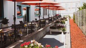An outdoor café with tables and chairs, red umbrellas, and potted plants. - Hotel Le Village (Winnenden)
