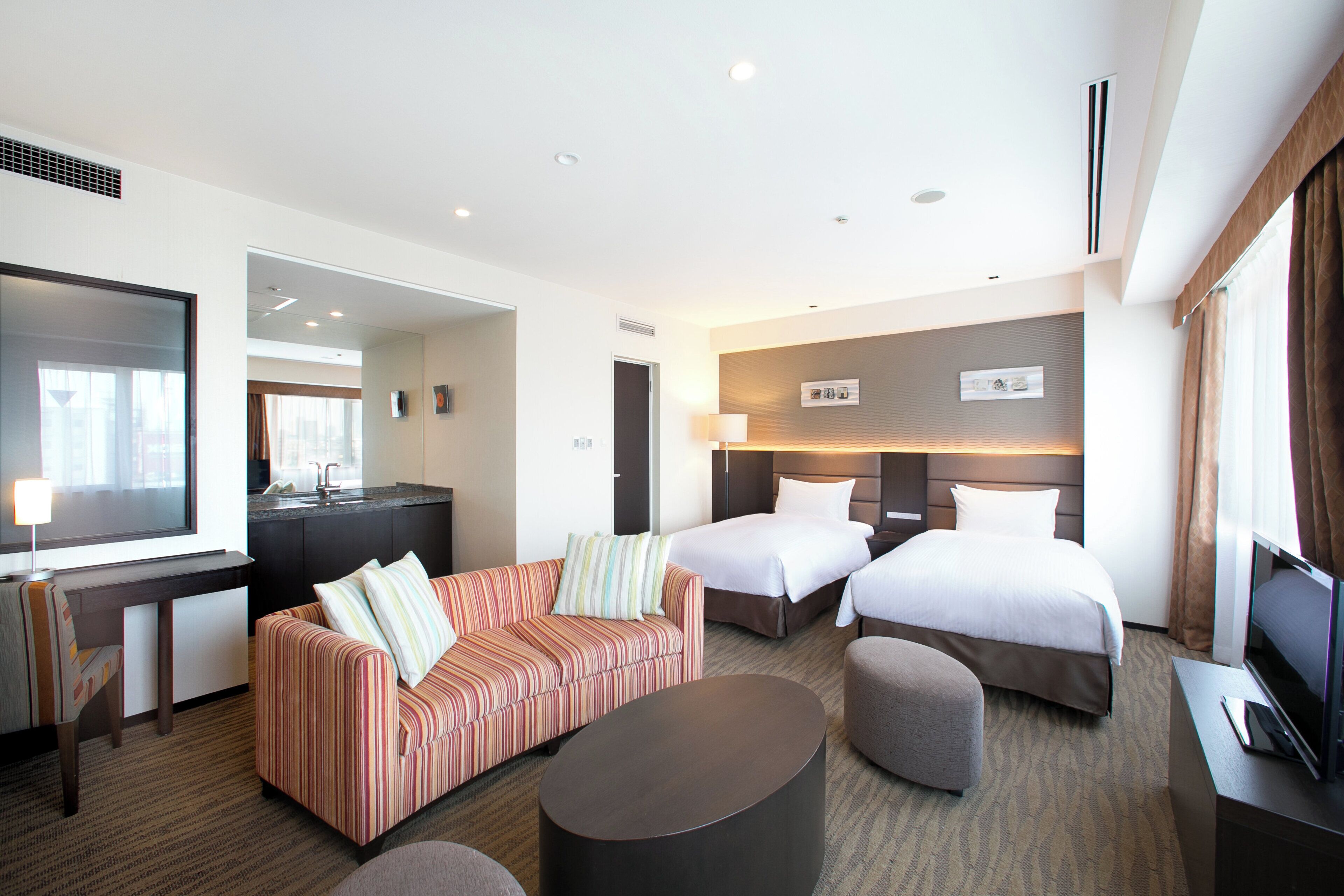 A hotel room with a sofa, two beds, a desk, a TV, and a bathroom.