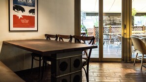 Een restaurantinterieur met een houten tafel en stoelen, een poster aan de muur en een uitzicht op het terras door een glazen deur.