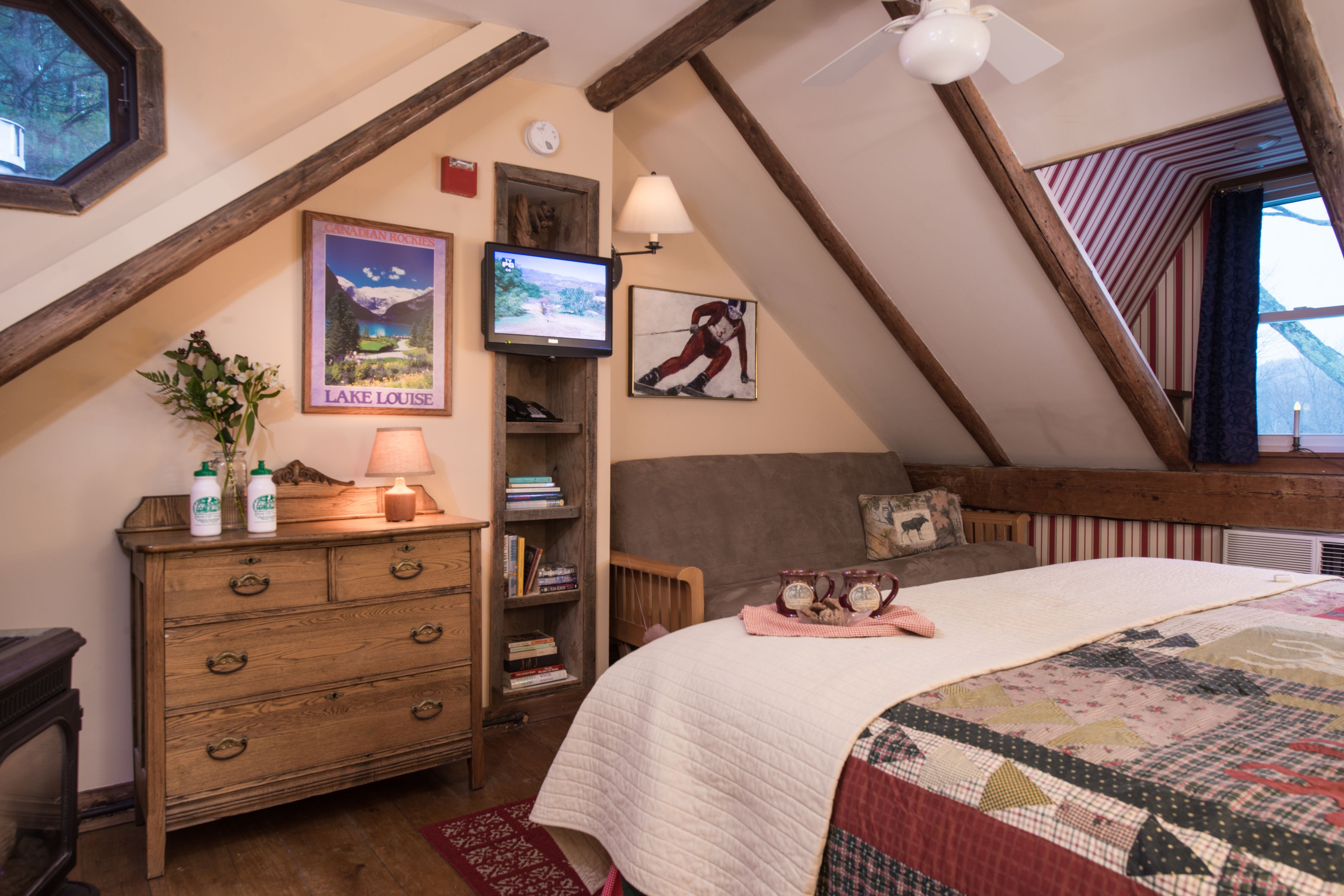 A cozy bedroom with a wooden dresser, a television, a bookshelf, and a large bed with a quilt.