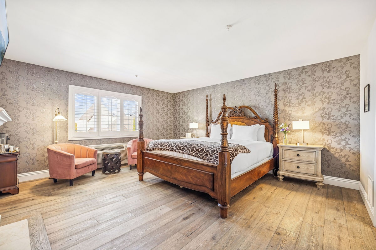 A four-poster bed with a wooden frame and a patterned bedspread, a pink armchair, a small round table, and a nightstand with a lamp.