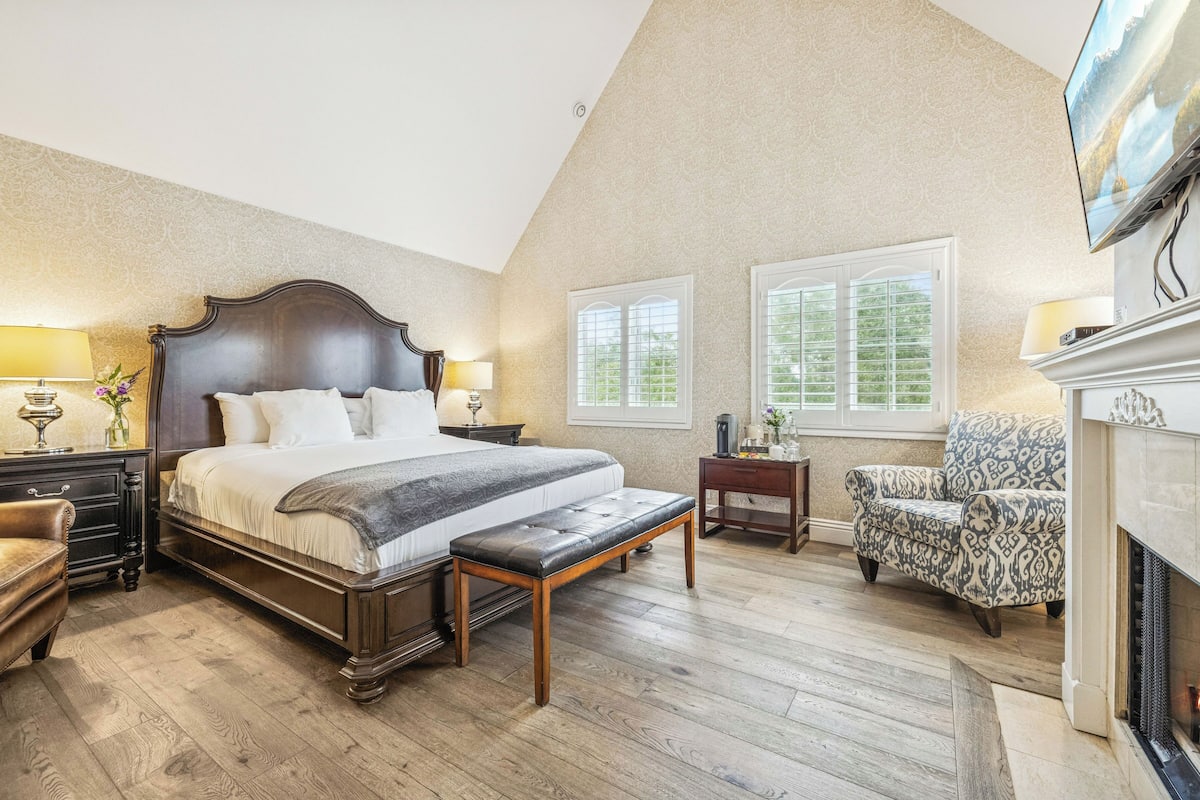 A bedroom with a large bed, bedside tables, a bench, a chair, a fireplace, and two windows.