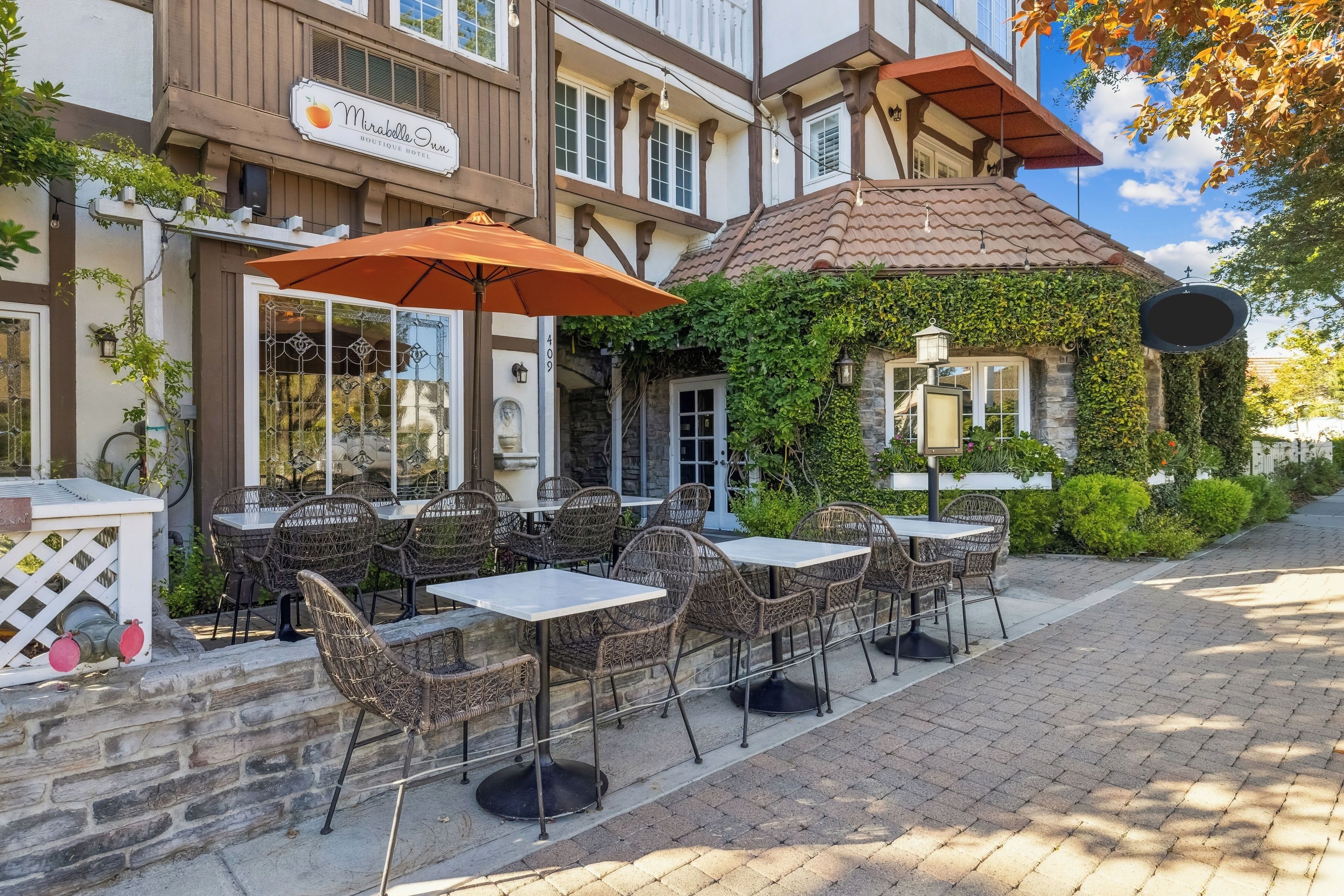 An outdoor cafe area with tables and chairs, a sign for Mirabelle Inn, and a building with ivy and stone walls.