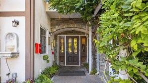 A stone archway leading to a dark wooden door with a doormat, surrounded by lush greenery and a brick pathway. - Mirabelle Inn (Solvang)