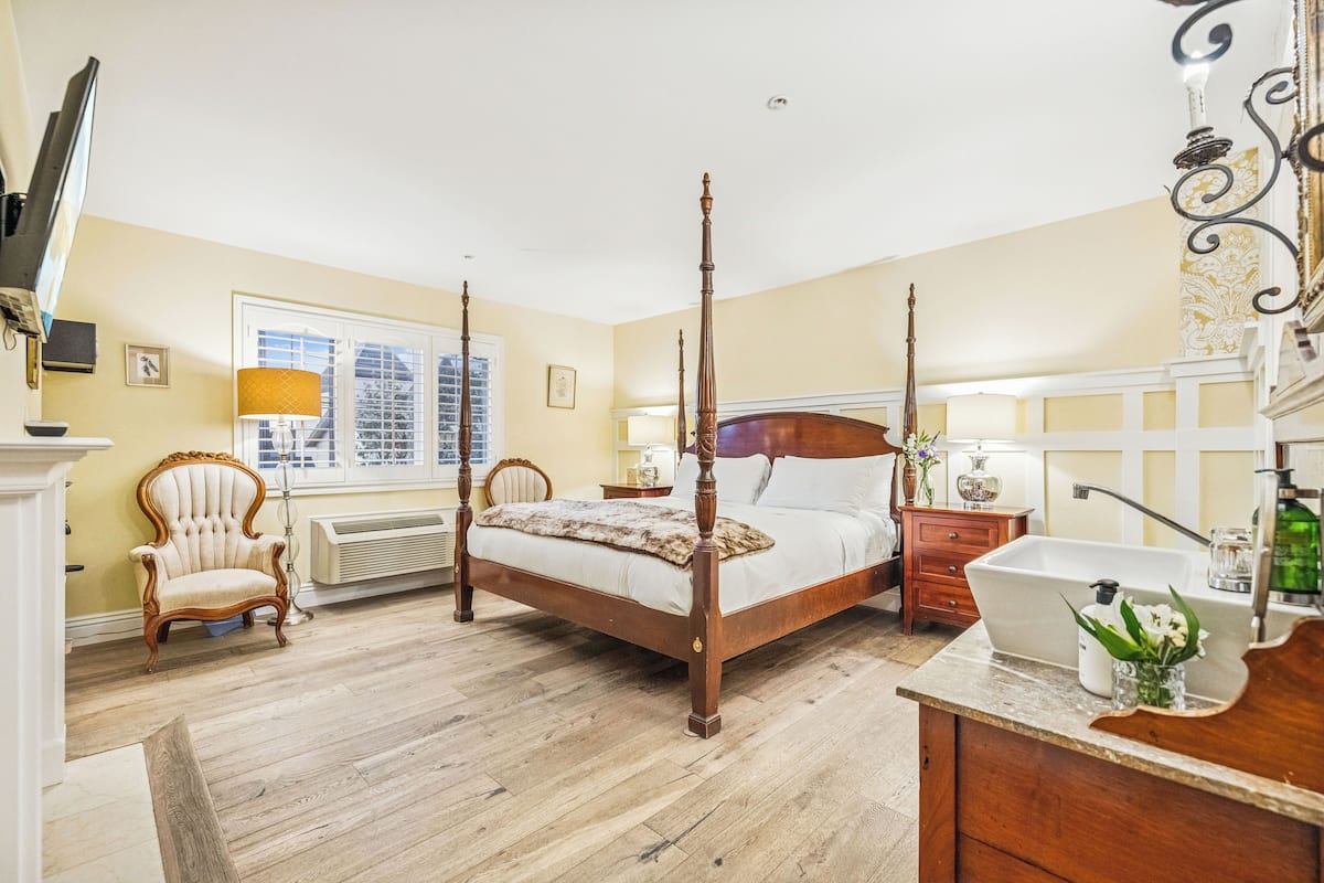 A four-poster bed with a wooden frame, a bedside table with a lamp, a chair, and a bathroom with a sink and mirror.