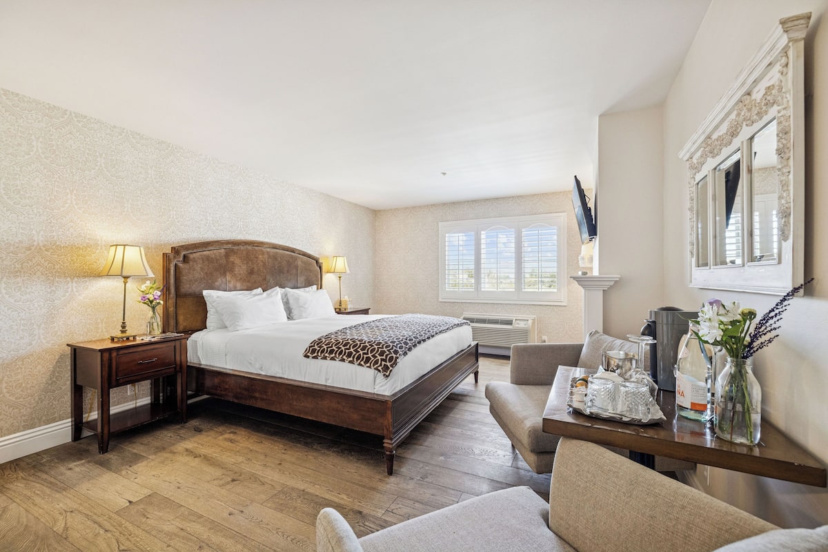 A hotel room with a large bed, a desk with a chair, a window, and a mirror on the wall.