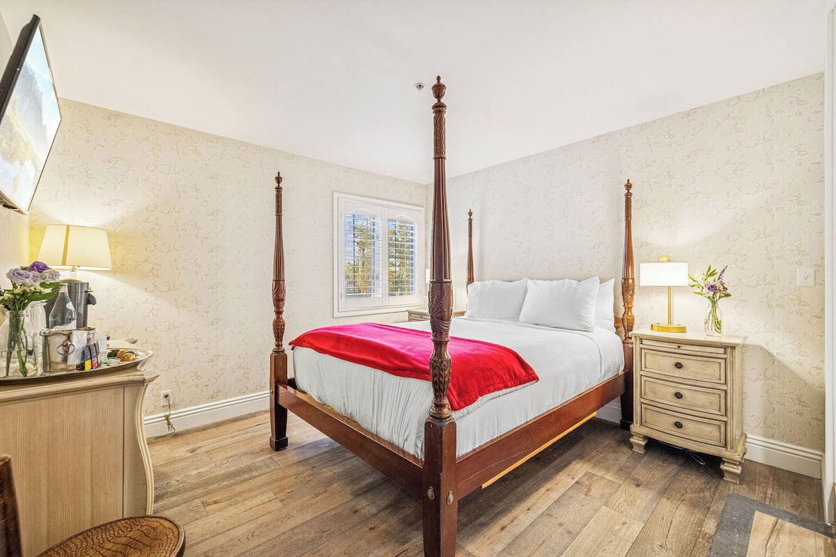 A four-poster bed with a red throw, a bedside table with a lamp, and a dresser with a mirror.