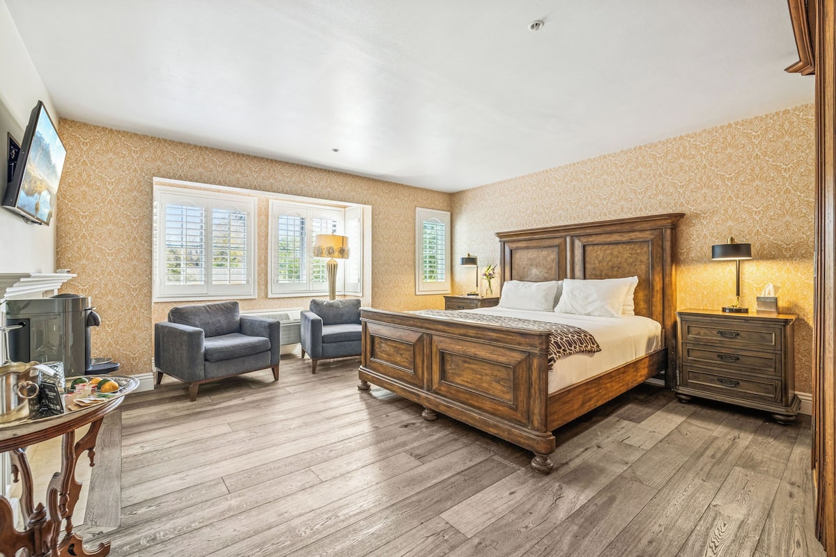 A bedroom with a large wooden bed, two armchairs, a dresser, and a table with a kettle and teapot.