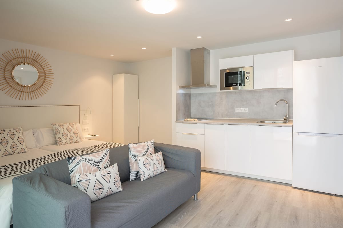 Un espacio habitable moderno y funcional con kitchenette, un sofá con cojines estampados y un espejo decorativo en la pared.
