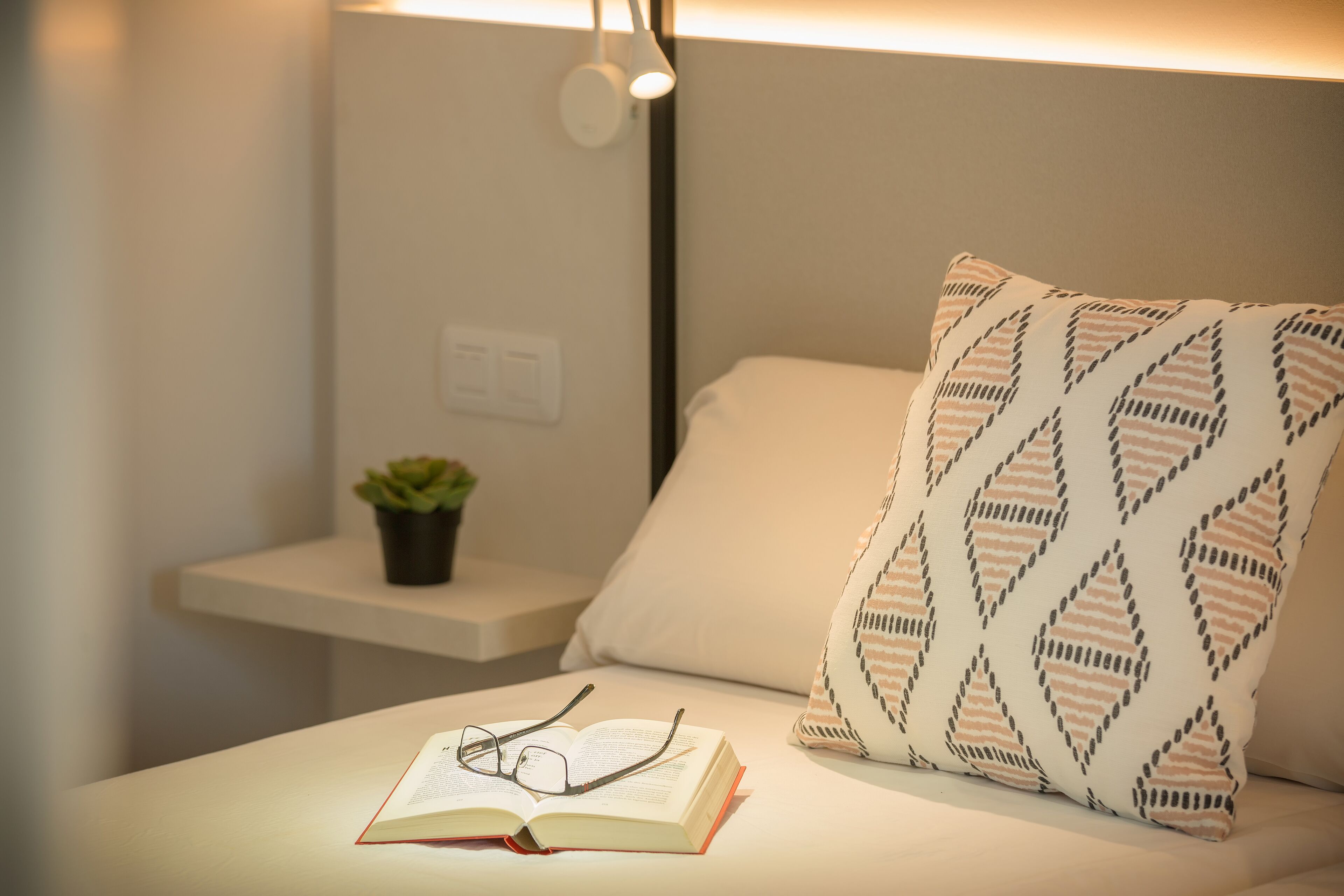 Una cama bien hecha con un almohadón estampado, una mesita de noche con una planta y un par de gafas sobre un libro abierto.