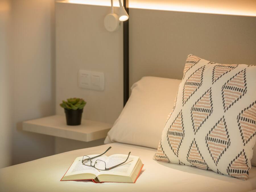 Una cama bien hecha con un almohadón estampado, una mesita de noche con una planta y un par de gafas sobre un libro abierto.