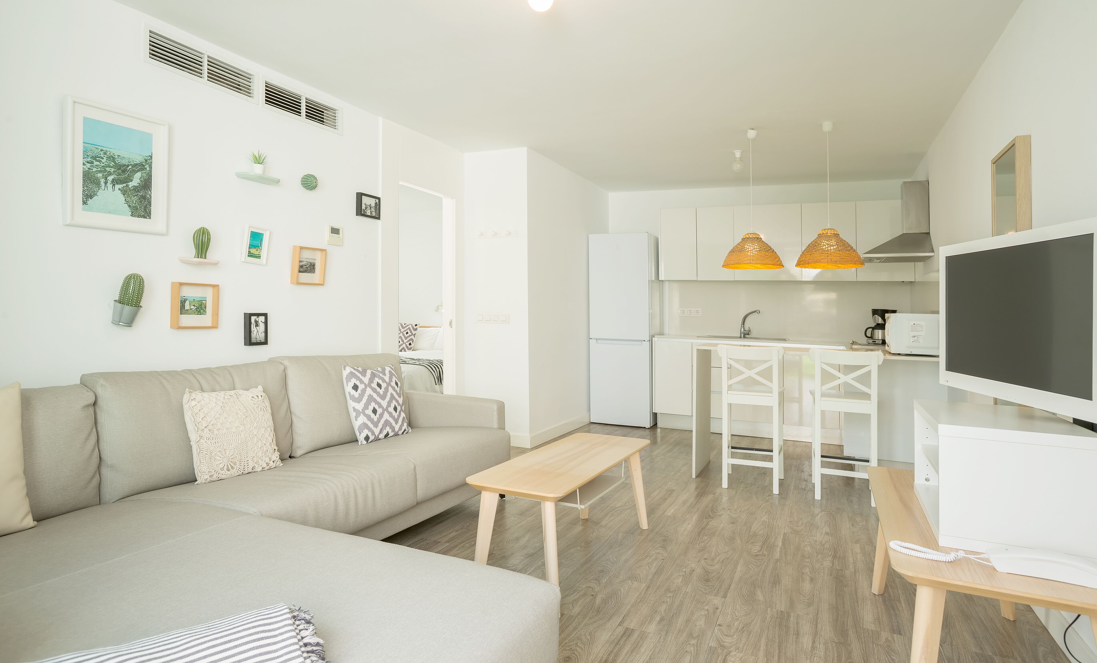 A modern living room with a sofa, a coffee table, and a TV. There is a kitchen area with a fridge, microwave, and a small dining table.