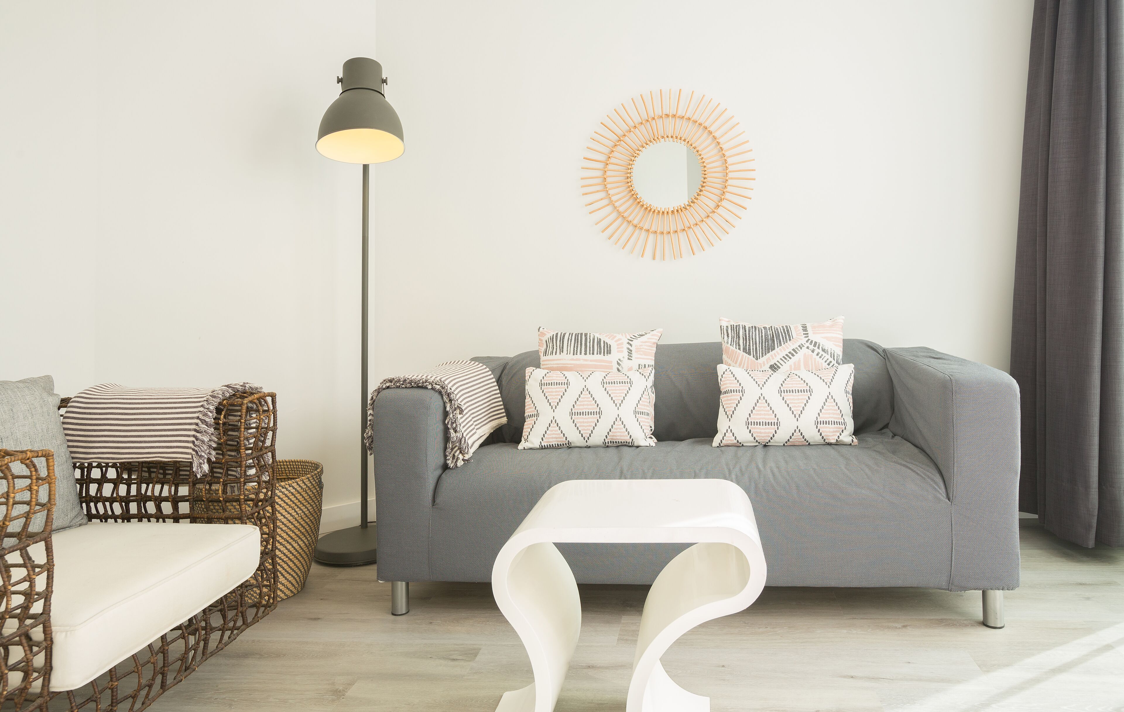 Una sala moderna con un sofá gris, cojines estampados, una silla de mimbre, una lámpara de pie y una mesa de centro blanca.