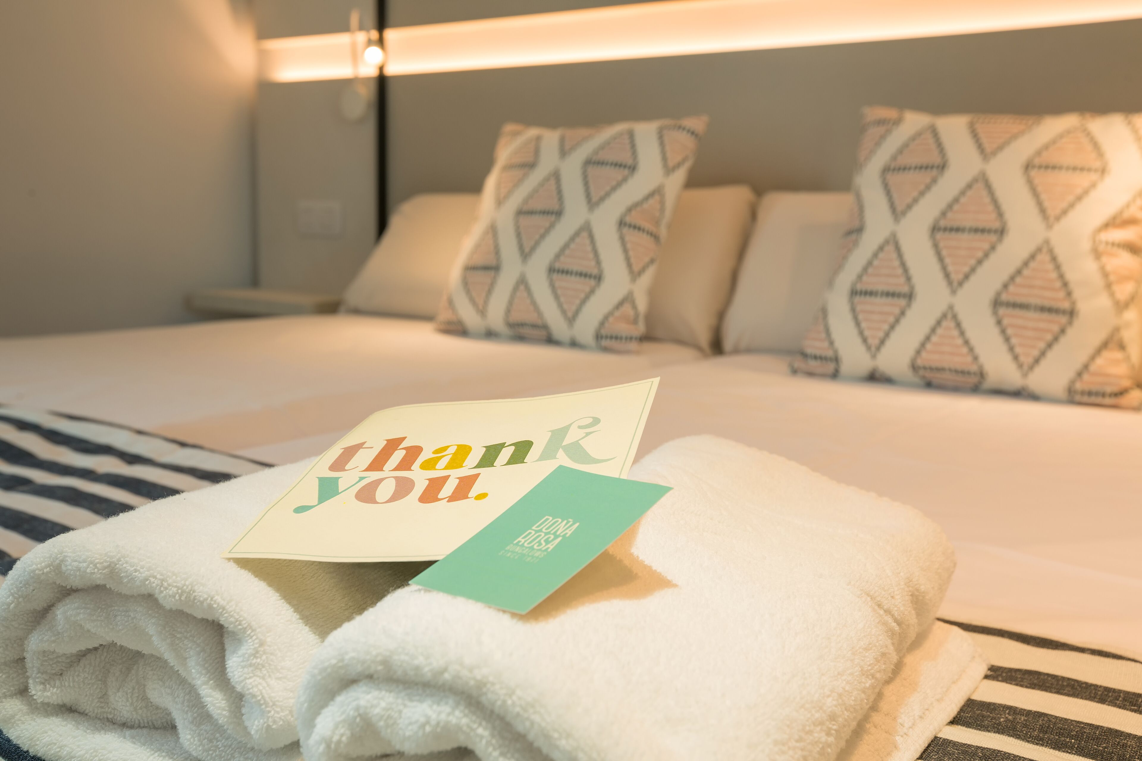A neatly made bed with a folded white towel and a thank you card on top.