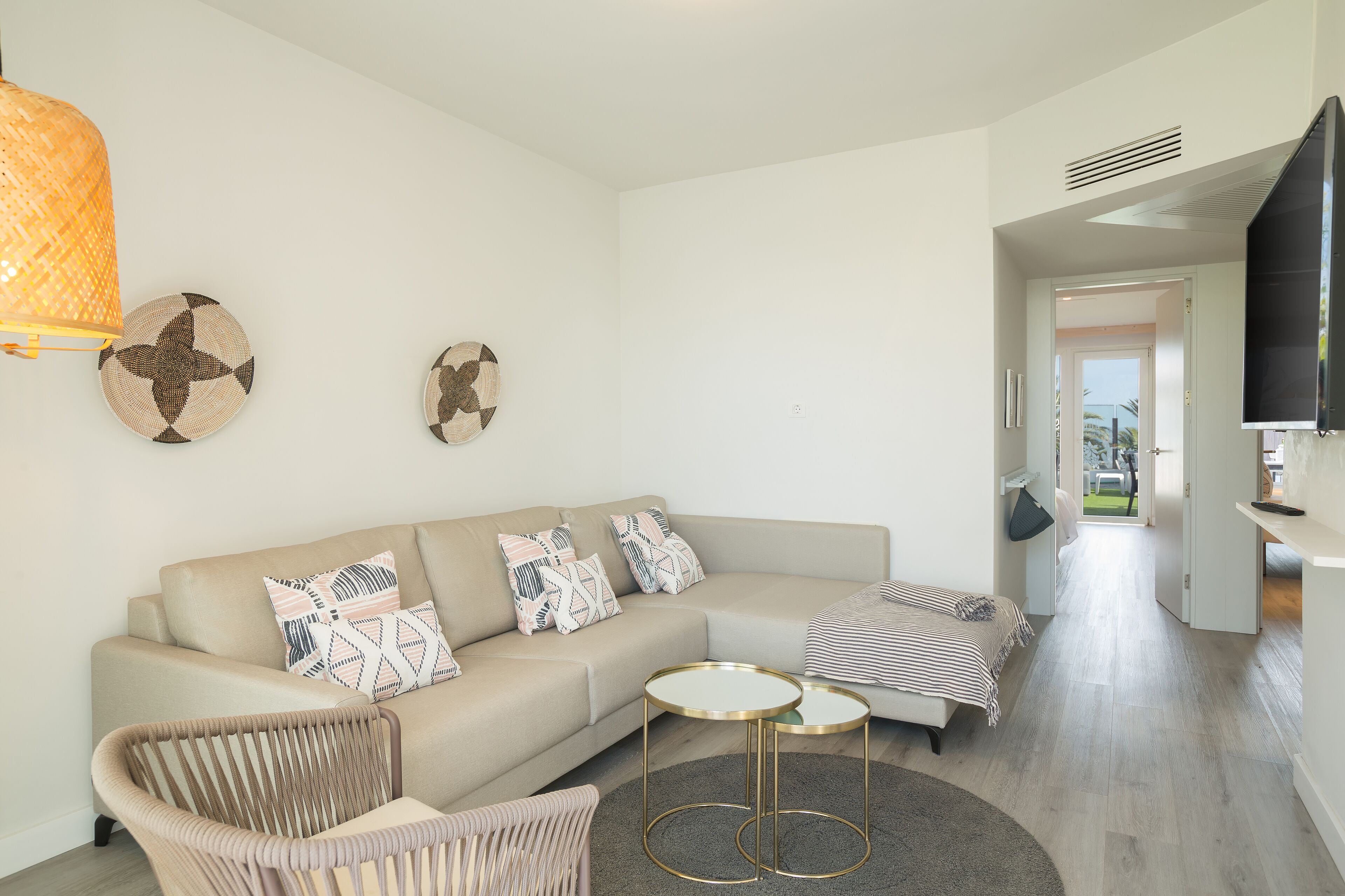 Un salon moderne avec un canapé beige, deux petites tables rondes, un fauteuil en osier et un téléviseur à écran plat fixé au mur.