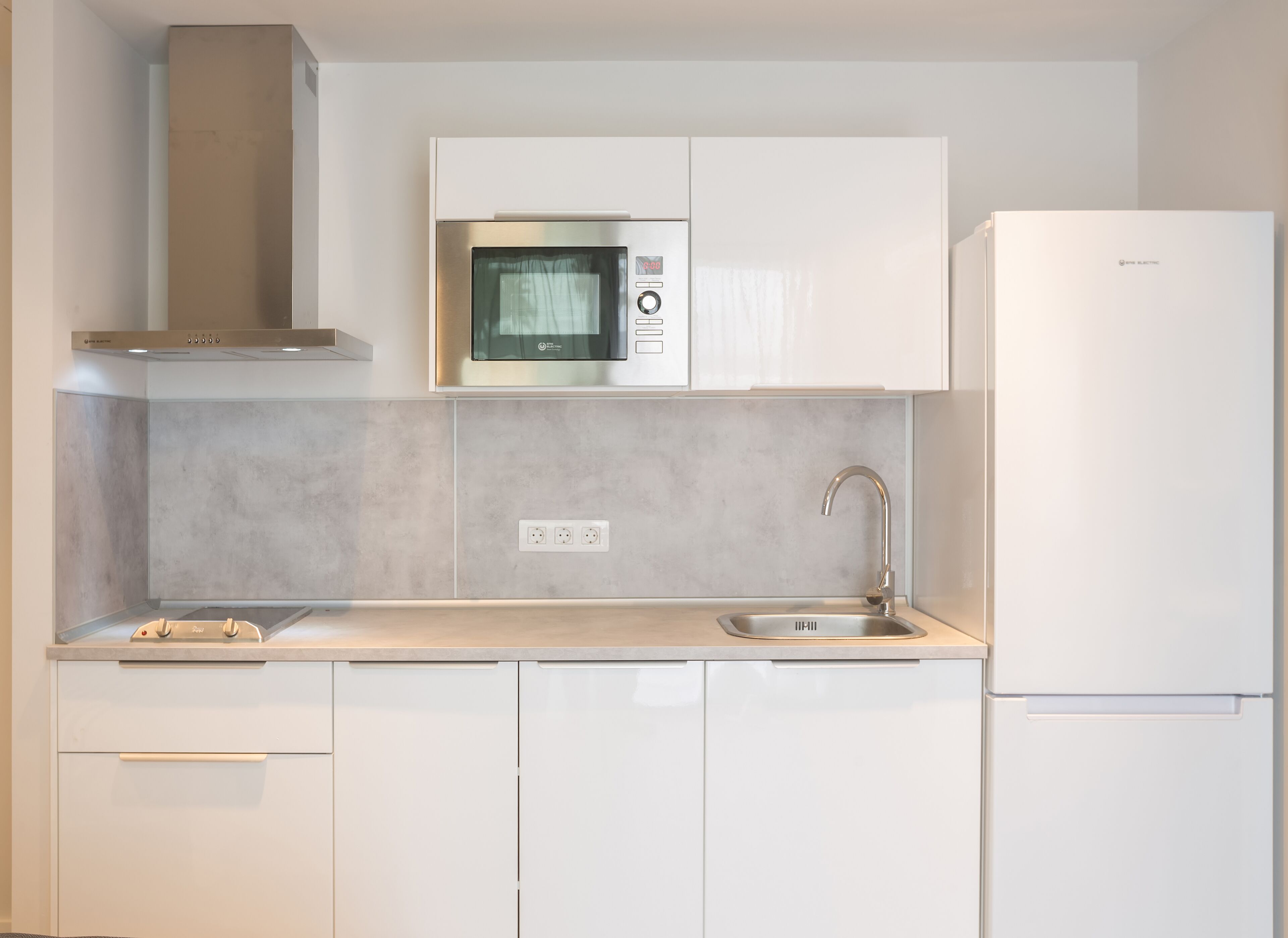 A modern kitchen with white cabinets, a stainless steel microwave, a built-in oven, a sink with a faucet, and a refrigerator.