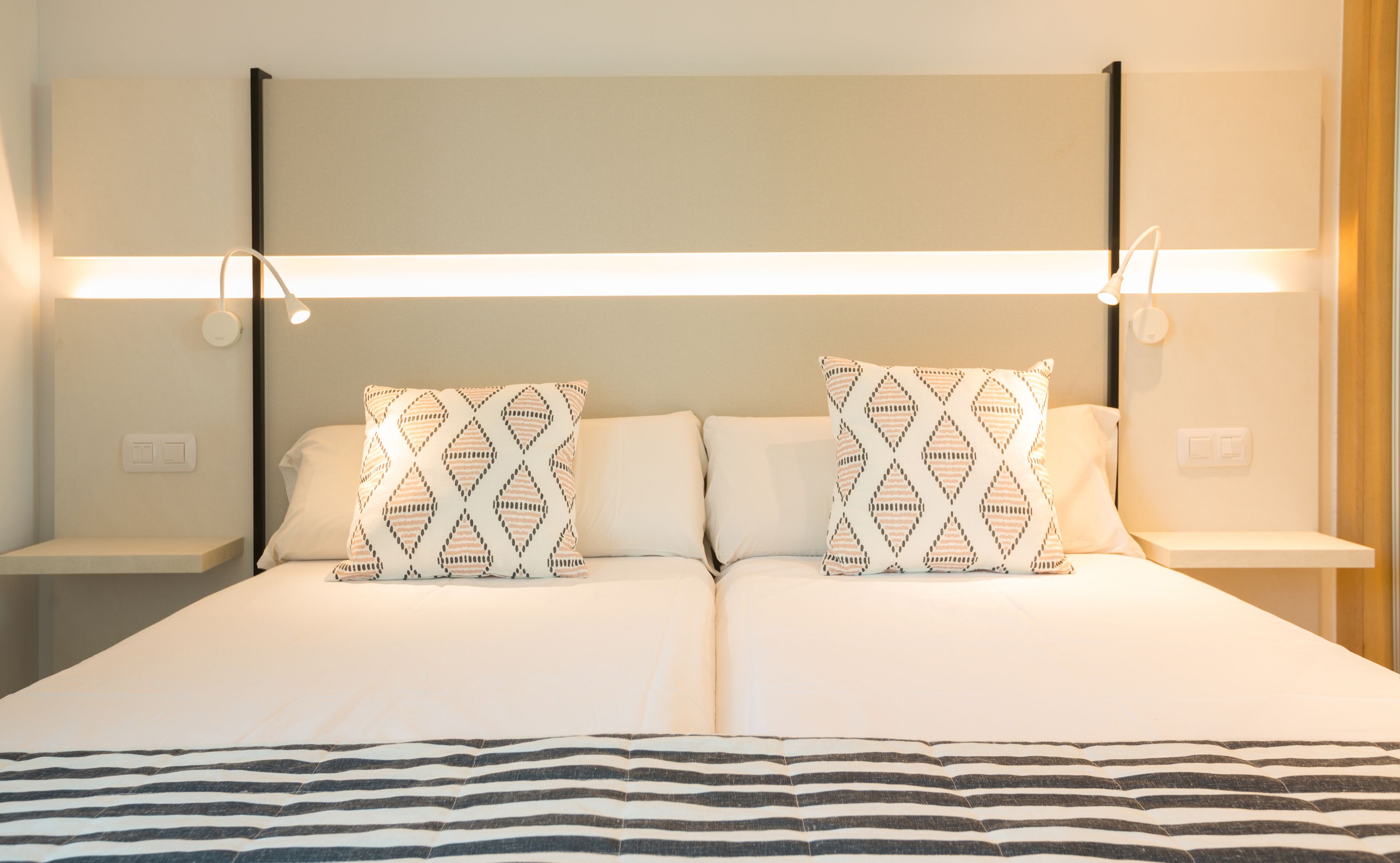 A double bed with two patterned pillows, a bedside table, and a wall-mounted light fixture.