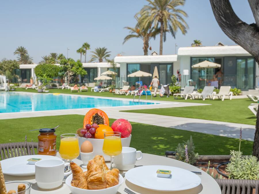 A breakfast setup with croissants, fruits, and orange juice on a table by a poolside with lounge chairs and palm trees in the background.