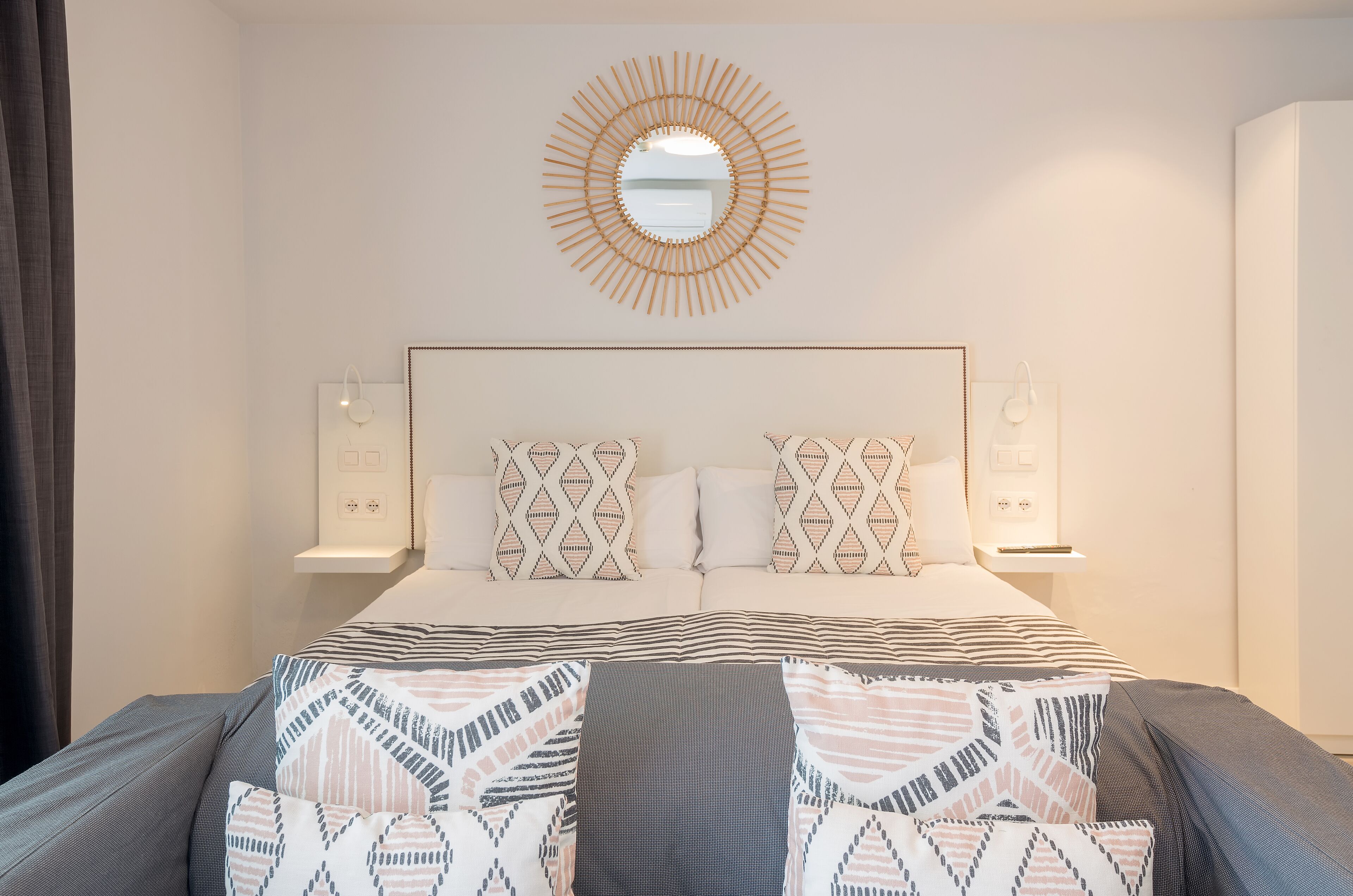 Ein modernes Schlafzimmer mit einem großen Bett, gemusterten Kissen, einem Wandspiegel, Nachttischlampen und einer neutralen Farbgestaltung.