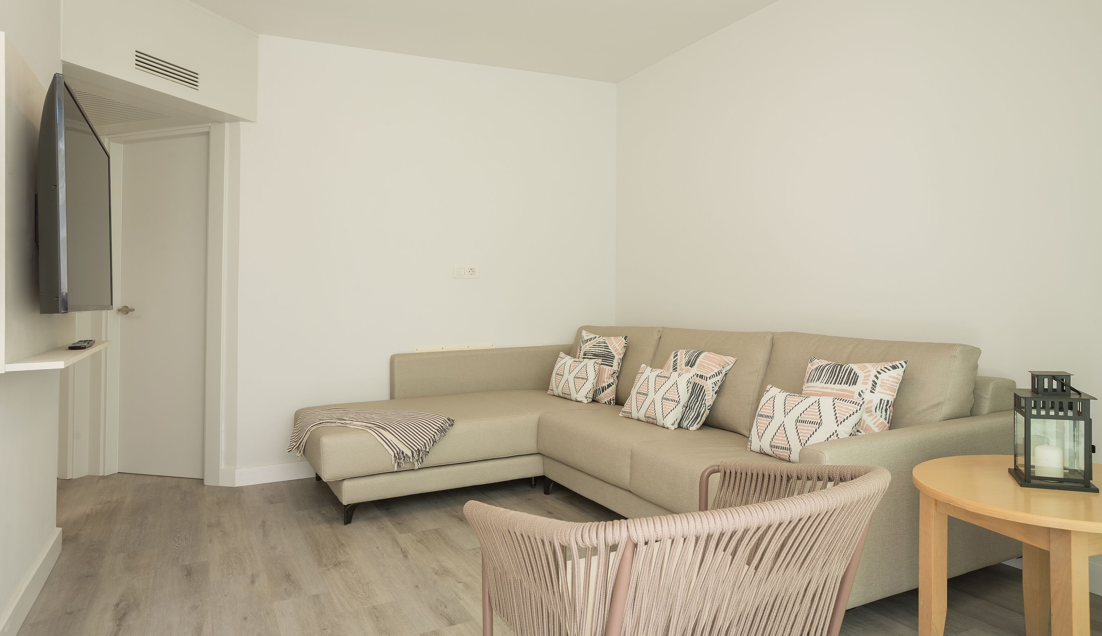 Ein modernes Wohnzimmer mit einem beigen Sofa, gemusterten Kissen, einem Korbstuhl, einem kleinen Holztisch und einem an der Wand befestigten Fernseher.