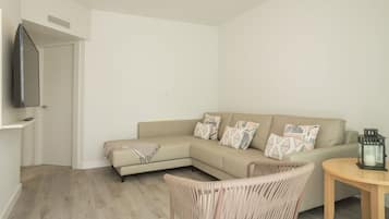 Ein modernes Wohnzimmer mit einem beigen Sofa, gemusterten Kissen, einem Korbstuhl, einem kleinen Holztisch und einem an der Wand befestigten Fernseher.