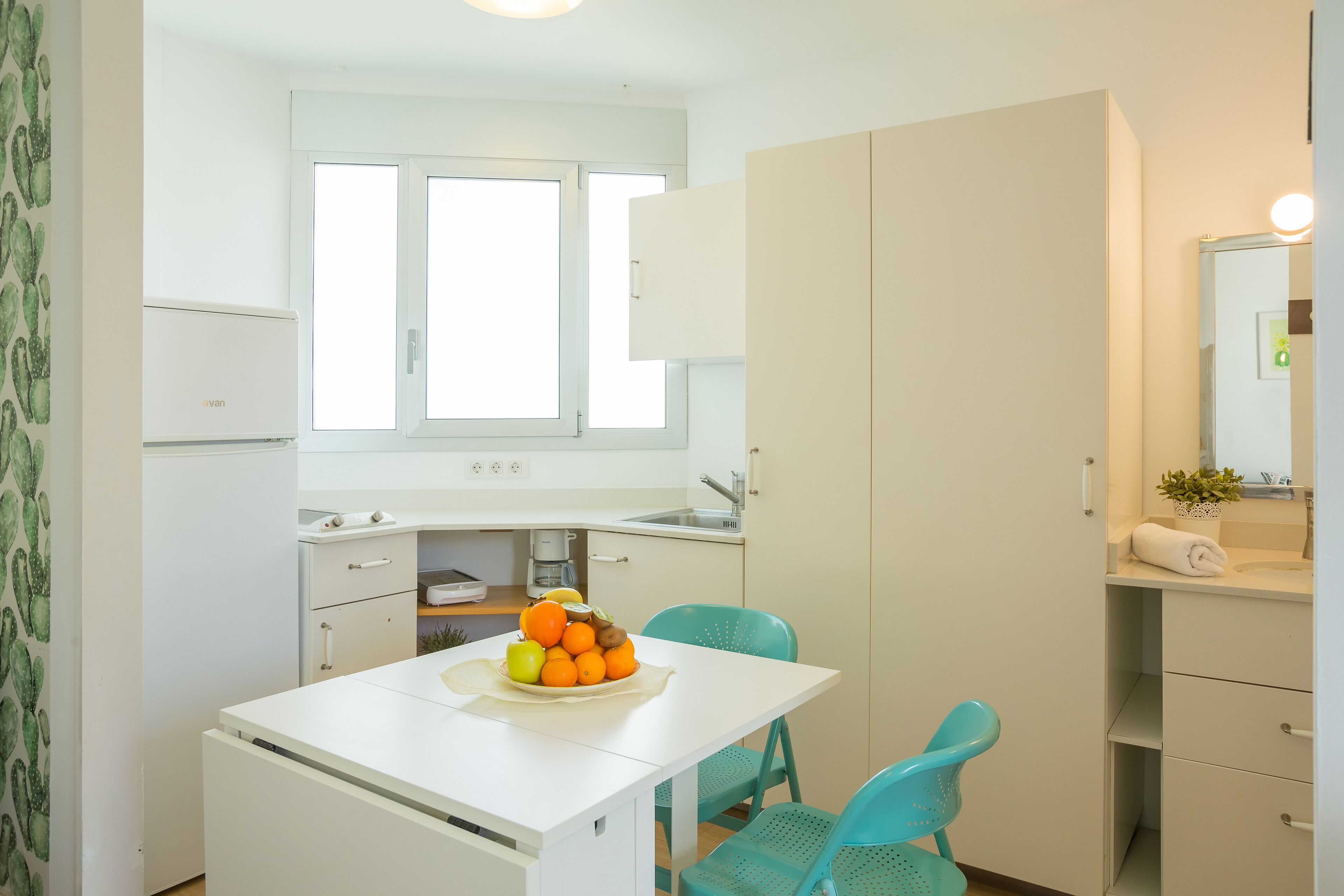 A compact kitchen with a white countertop, a sink, and a window above the counter. There is a small table with a fruit bowl and a teal chair.