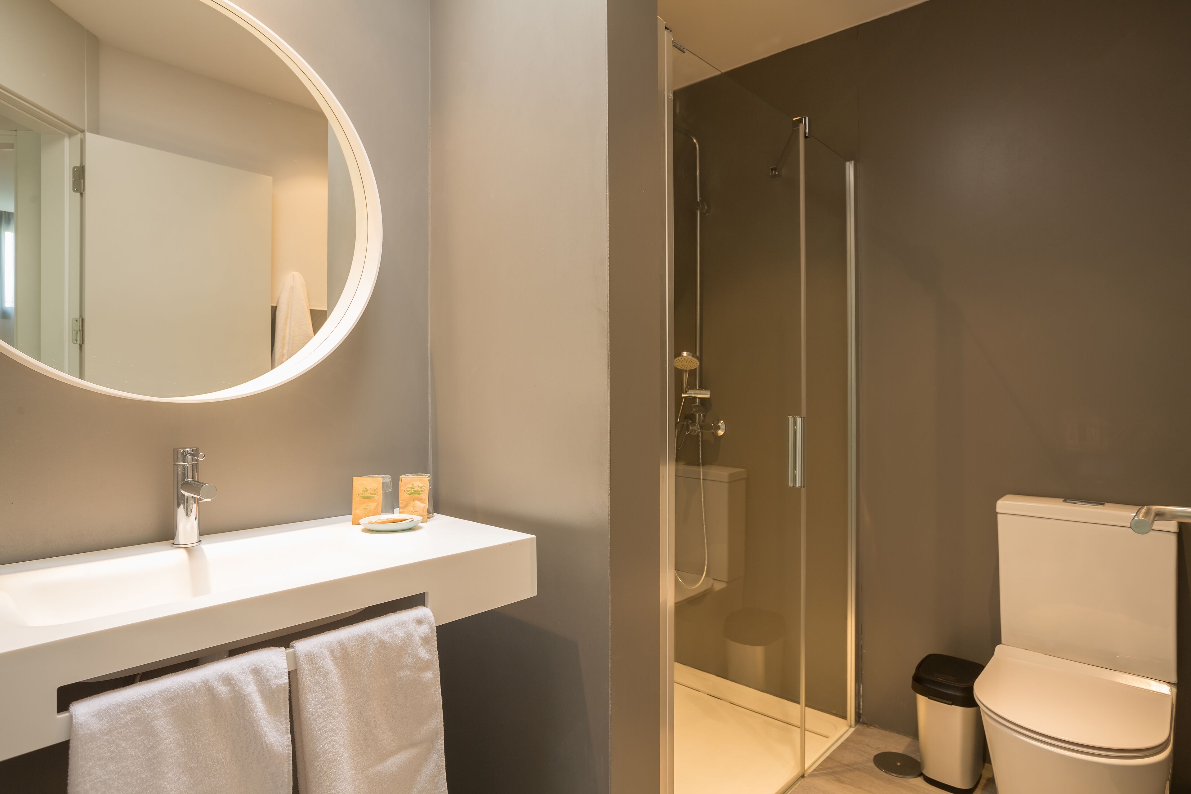 A modern bathroom with a white sink, a round mirror, a glass-enclosed shower, and a toilet.