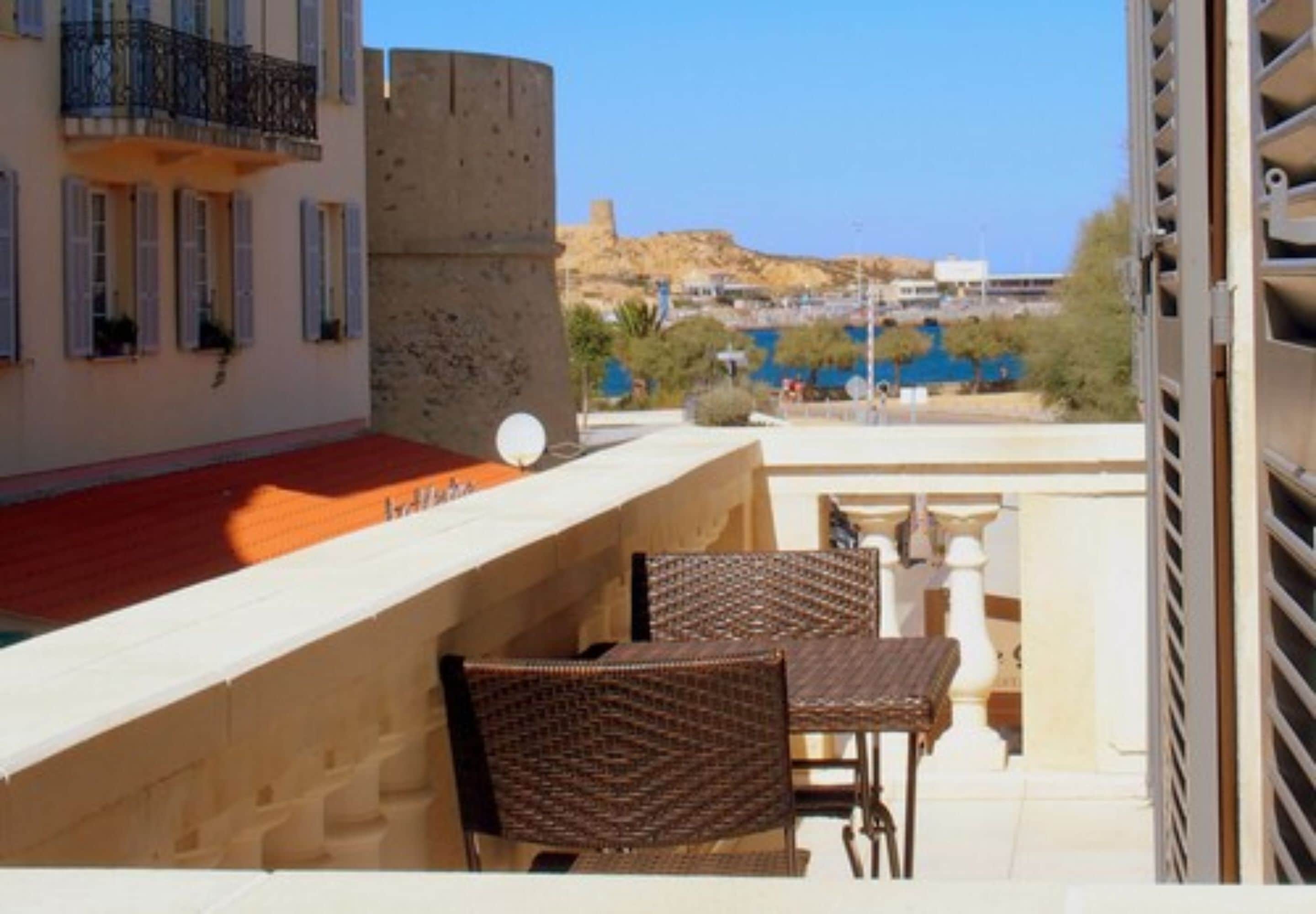 Un balcone con sedie in vimini e un tavolo, con vista su una zona costiera con edifici e una collina.