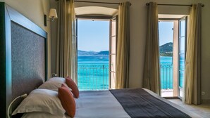 A bedroom with a bed, pillows, a lamp, and a view of the sea through open curtains. - Hôtel Perla Rossa (L'Île-Rousse)