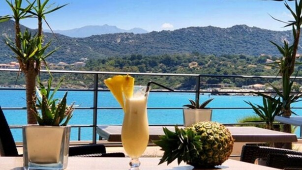 A table with a drink, a pineapple, and a potted plant, overlooking a body of water and mountains. - Hôtel Perla Rossa (L'Île-Rousse)