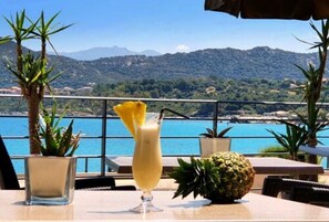 A table with a drink, a pineapple, and a potted plant, overlooking a body of water and mountains. - Hôtel Perla Rossa (L'Île-Rousse)