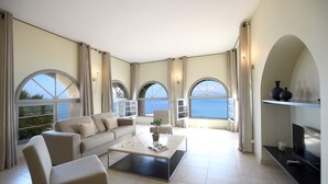 A spacious living room with a sofa, armchairs, a coffee table, and a TV. Large windows offer a view of the sea. - Hôtel Perla Rossa (L'Île-Rousse)
