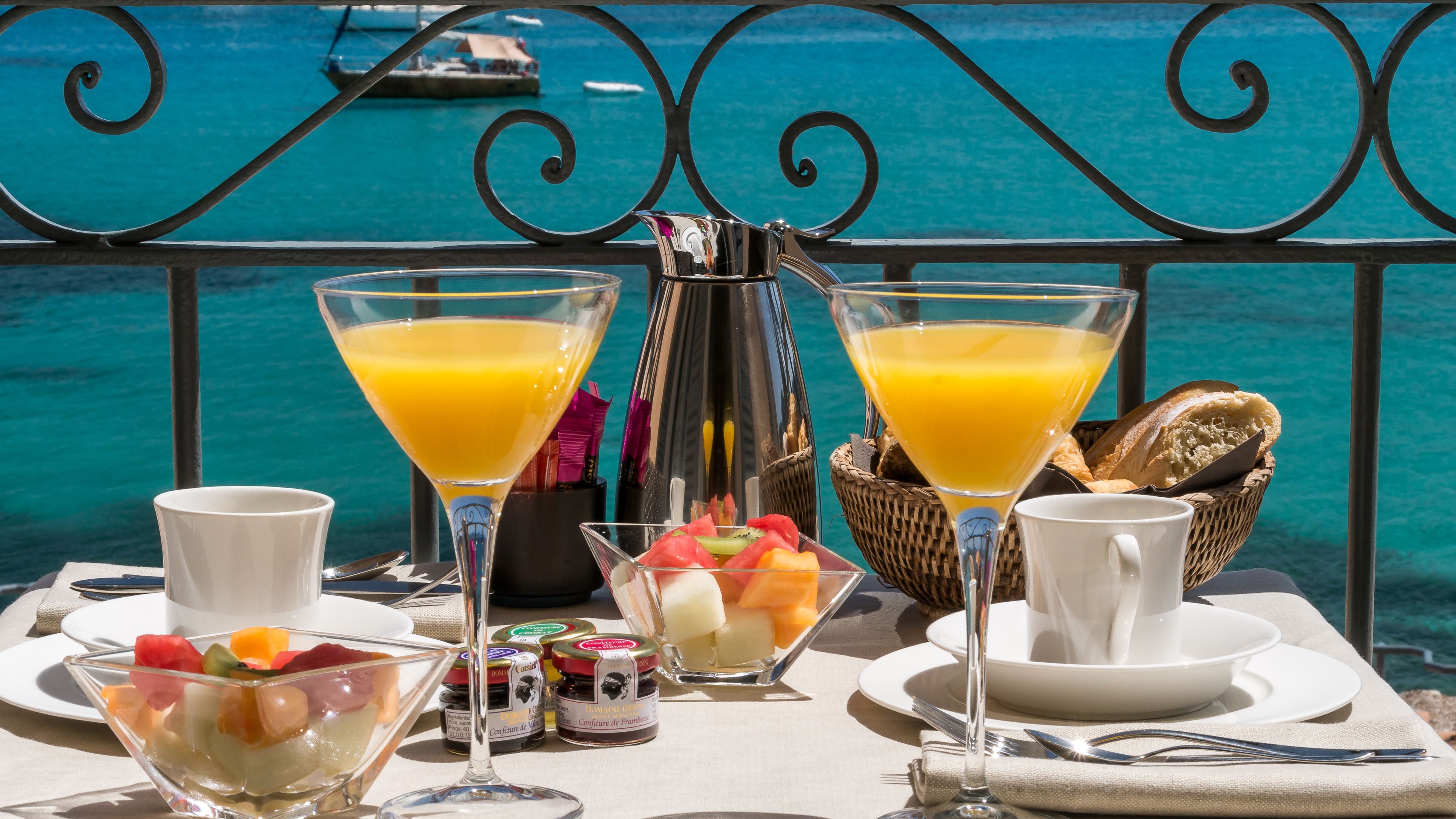 Un plateau de petit-déjeuner comprenant deux verres à martini remplis de jus d’orange, un saladier de fruits, un panier de pain et divers condiments, le tout disposé sur une table offrant une vue sur la mer.