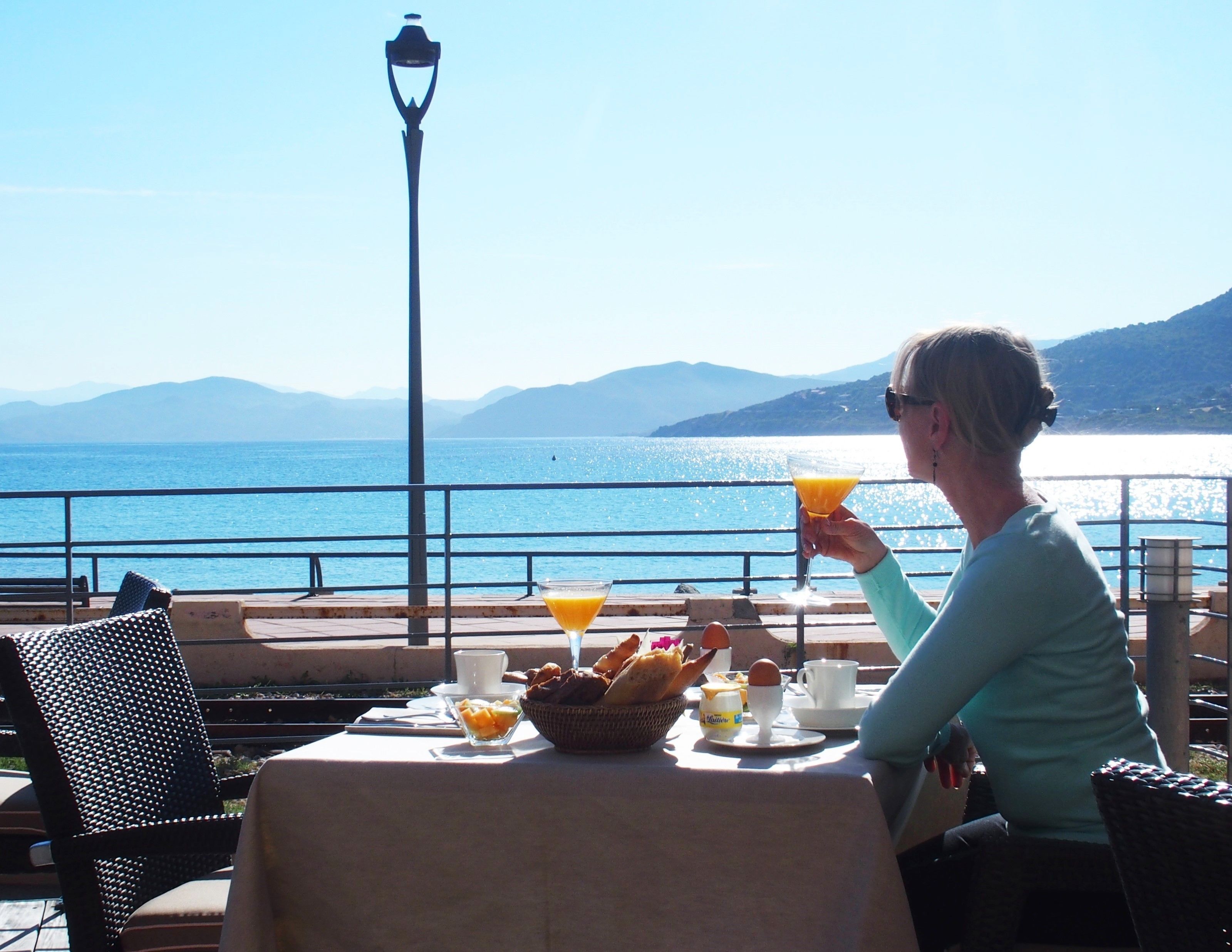 A table with breakfast items and a drink, overlooking a body of water with mountains in the background.