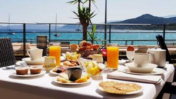 A breakfast table with various food items and drinks, overlooking a scenic coastal view with boats and mountains.
