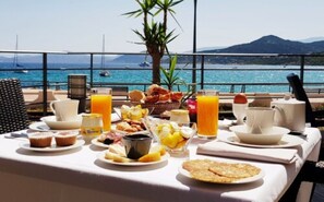 A breakfast table with various food items and drinks, overlooking a scenic coastal view with boats and mountains.