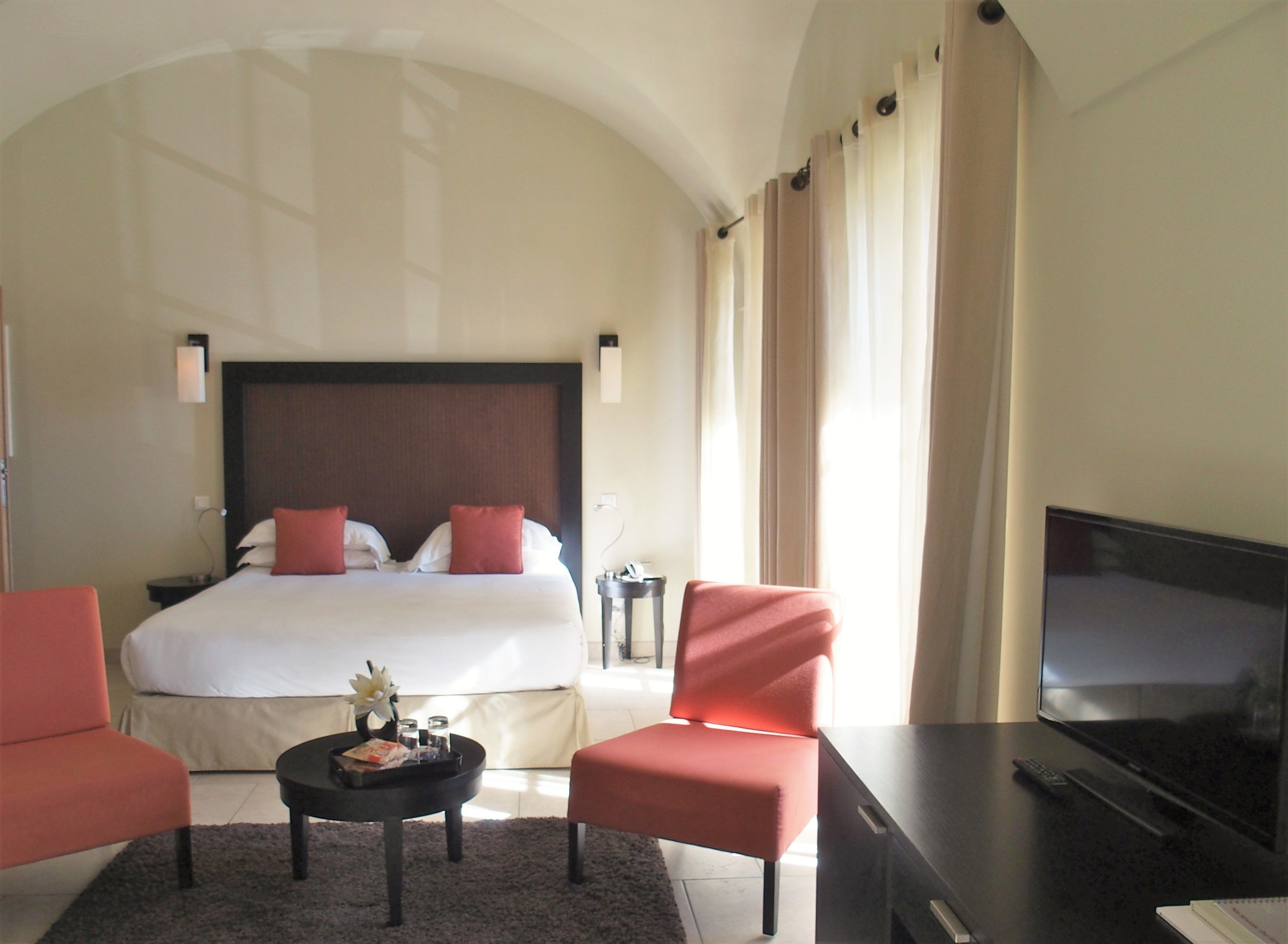 A hotel room with a large bed, two red chairs, a TV, a small table with a tray, and a nightstand with a lamp.