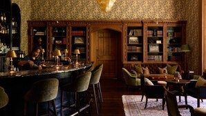 Uma área de bar com um bartender, uma estante de madeira e um espaço de estar com cadeiras e um sofá.