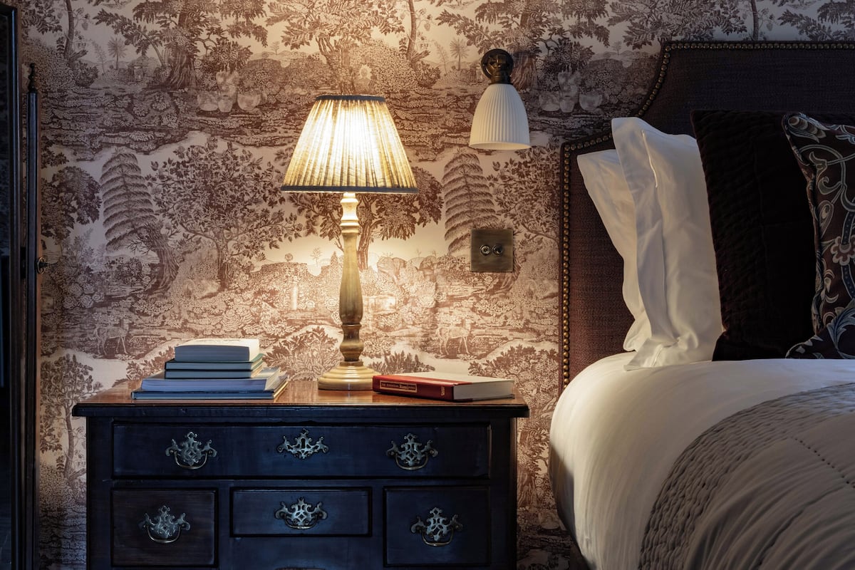 A neatly made bed with white linens, a bedside lamp, and a stack of books on a dark wooden nightstand.