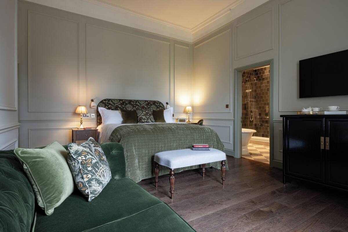 A hotel room with a green upholstered sofa, a matching bench, two bedside tables with lamps, a large bed with a patterned headboard, a white bathtub, and a black cabinet.