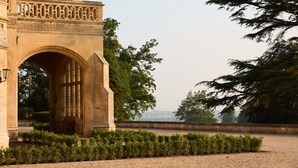 Um arco de pedra com um banco, cercado por sebes aparadas e árvores.
