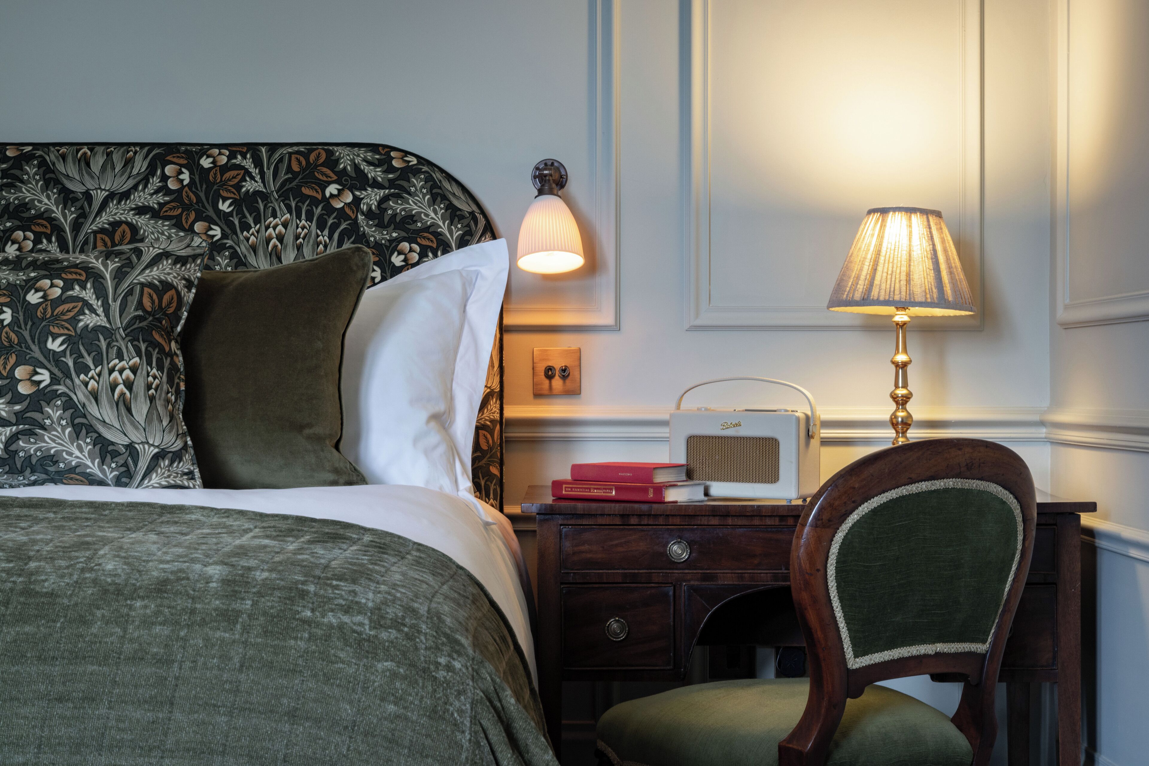A neatly made bed with a floral headboard, a bedside lamp, a radio, and a chair with a green cushion.