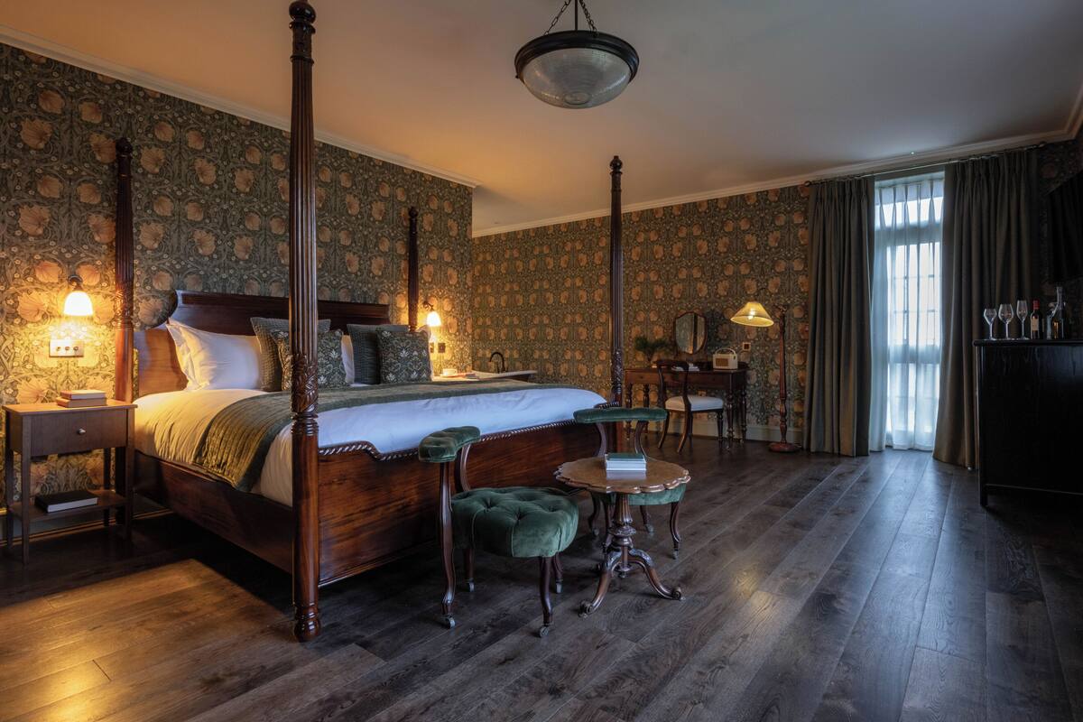 A four-poster bed with a wooden frame, a green upholstered footboard, and a matching headboard. There is a small round table with a green upholstered chair in front of the bed. The room has wooden flooring and a large window with curtains.