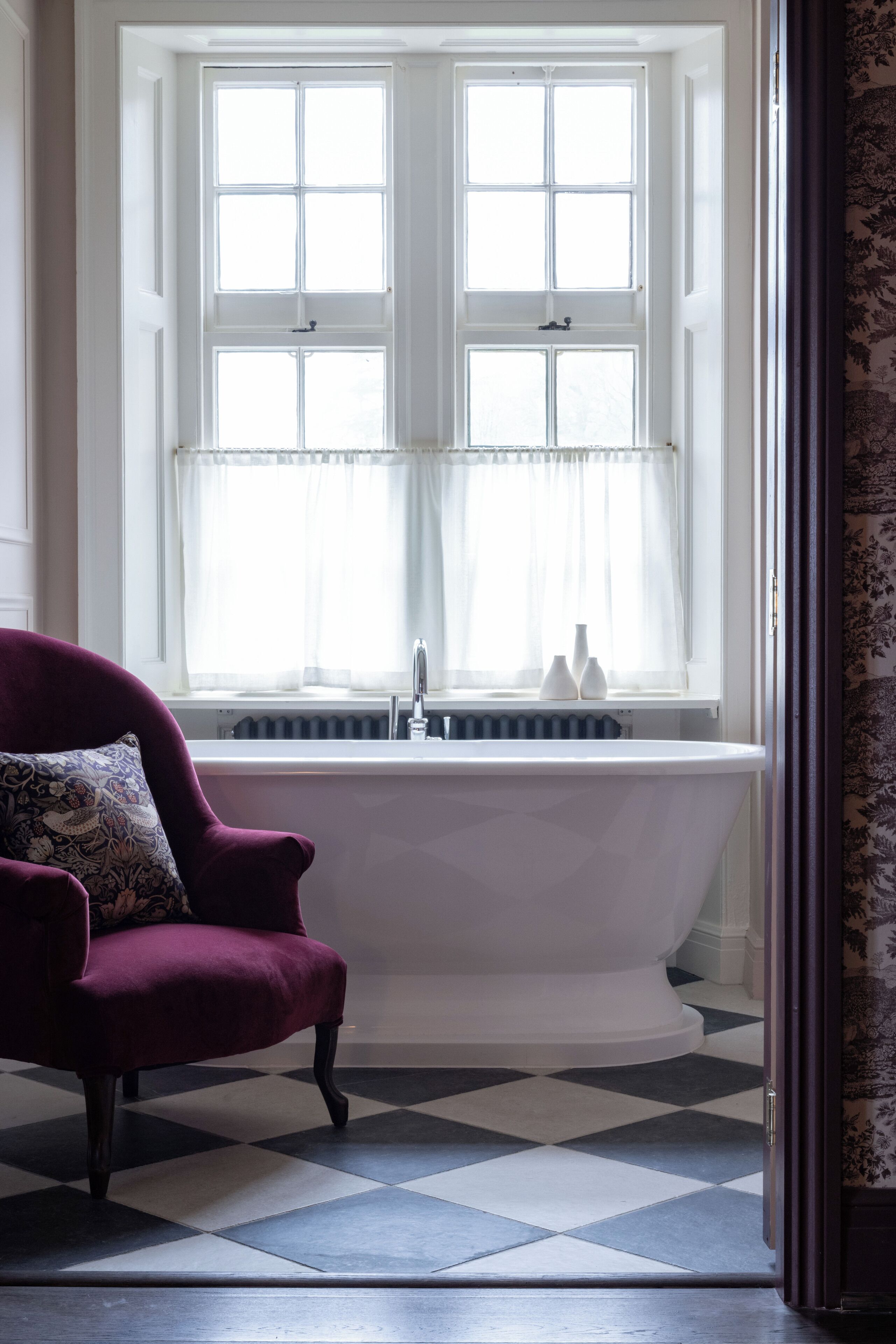 Une salle de bain avec une baignoire sur pieds, une fenêtre avec des rideaux blancs et un carrelage à carreaux.